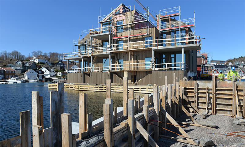 Trebygning under oppføring på kaifront med stillas, blå himmel og arbeidere i gule vester