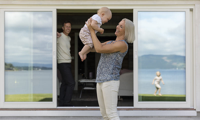 Kvinne i lys topp holder baby ved skyvedør, innsjø i bakgrunnen og barn leker ute
