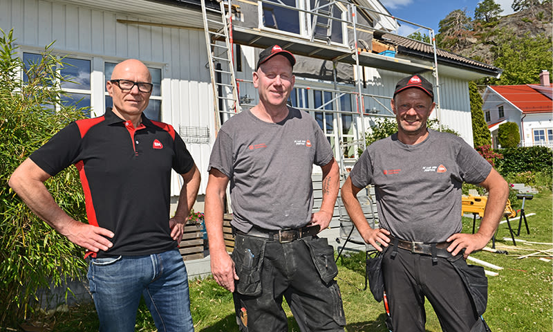 Tre håndverkere i grå og svart uniform foran hvitt hus med stillas og verktøy i hagen.