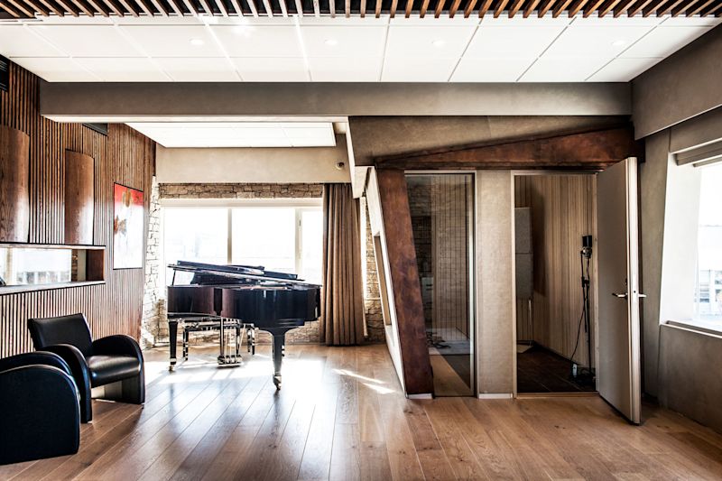 Moderne musikkstudio med svart flygel på lyst eikegulv, mørke trepaneler, lærstoler og vokalbås
