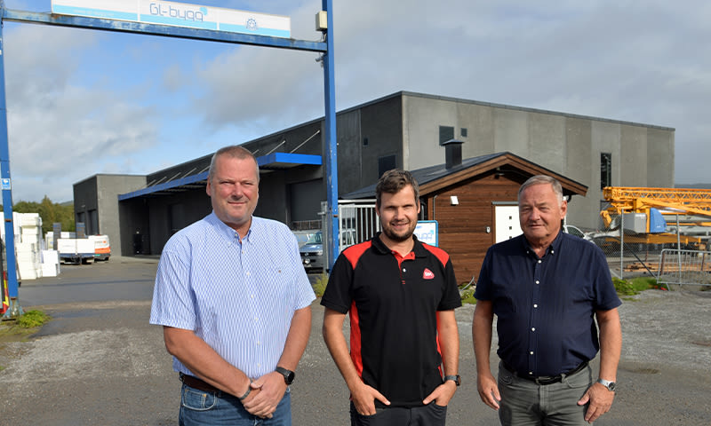 Tre menn på grusplan foran et grått fabrikkbygg med blå portal og en gul kran i bakgrunnen.