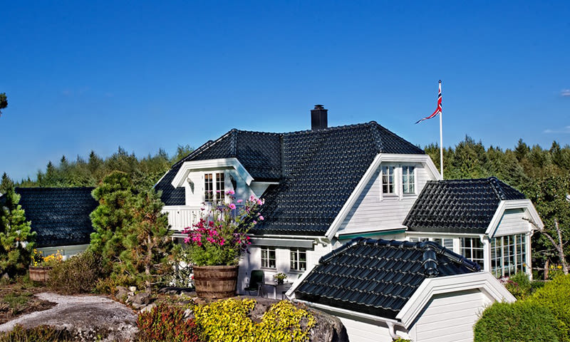 Hvit trevilla med svart tak, blomster i tønne, norsk flagg på stang, blå himmel