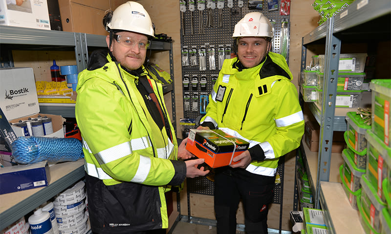 To arbeidere i gule vernejakker og hvite hjelmer i byggelager, holder oransje eske.