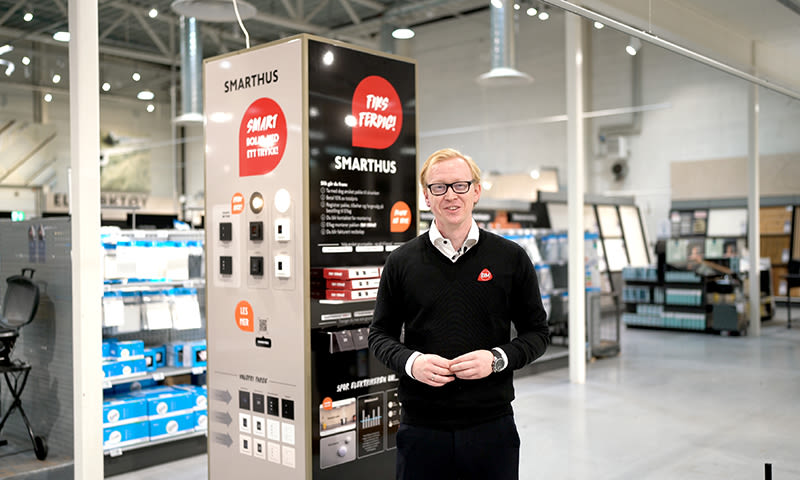 Butikkansatt i svart genser foran beige og svart smarthus-display med røde detaljer i lyst varehus