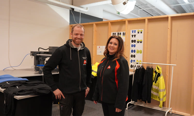 To ansatte i svarte arbeidsjakker foran klesstativ med gule refleksjakker.