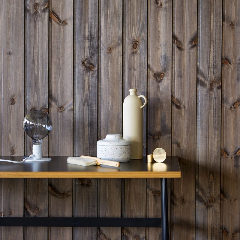 Mørk trevegg med svart bord, glasslampe, hvit keramikkkrukke, beige mugge, pensel og gullobjekter.