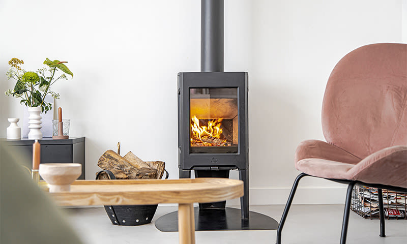 Moderne stue med svart vedovn som brenner, rosa velurstol, lyst bord, vedkurv og grønne planter.