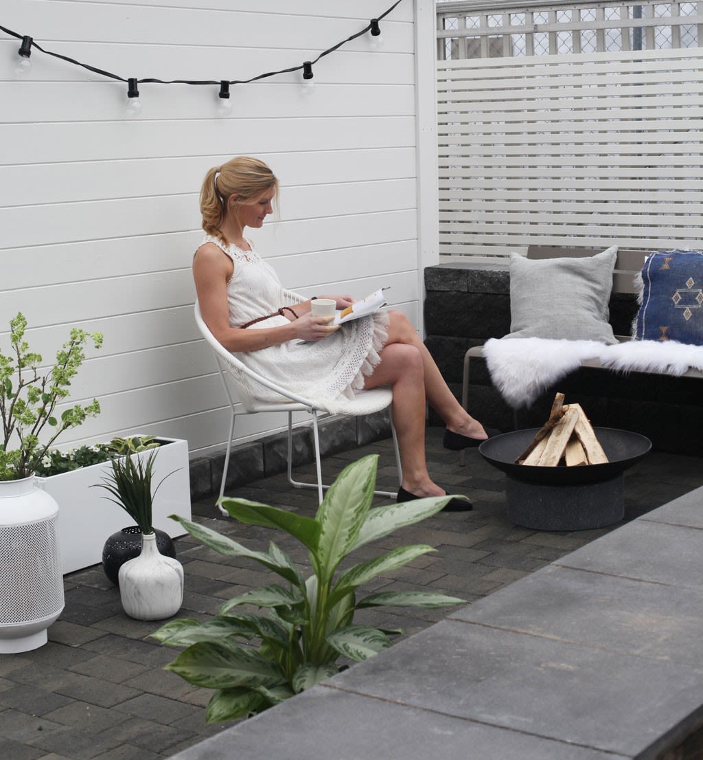 Kvinne i hvit kjole leser og drikker kaffe på grå-hvit uteplass med planter og bålfat