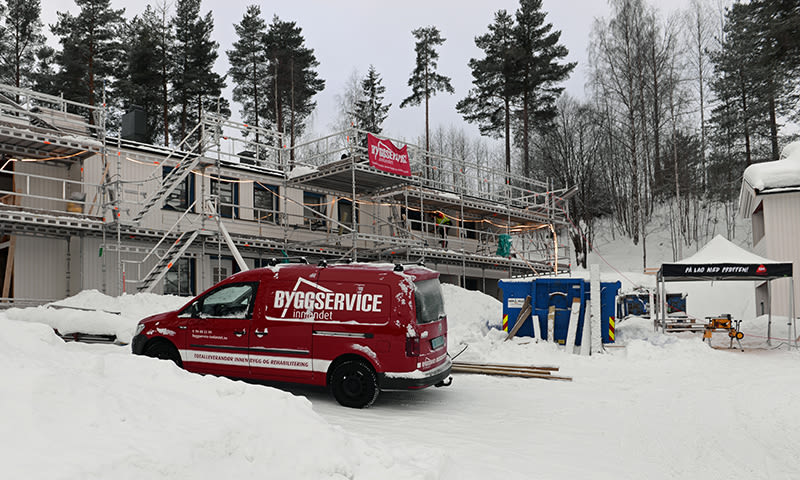 Snødekt anleggsplass med hvit bygning på stillas og rød Byggservice-bil i skog.