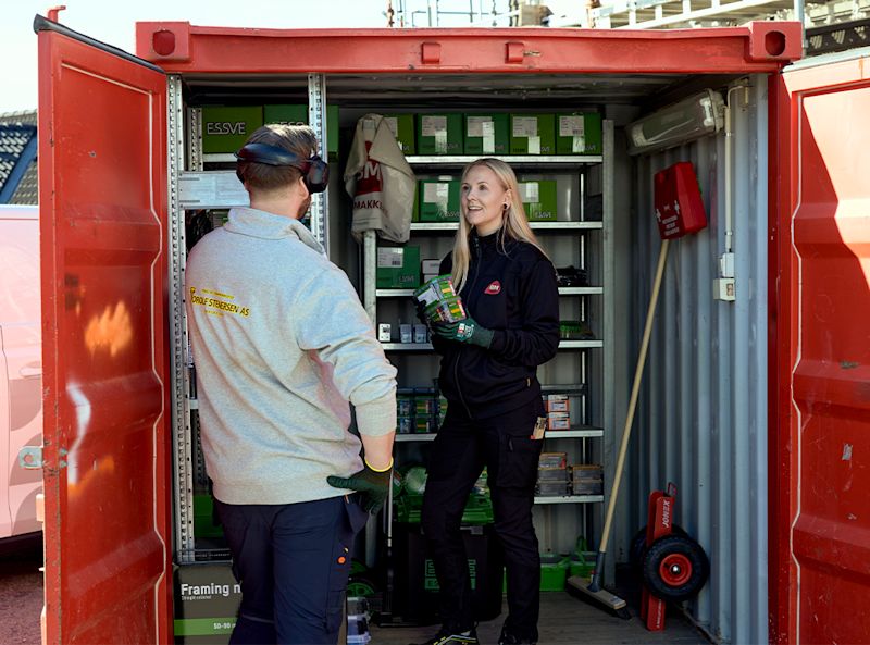 Rød container med hyller av grønne esker, to arbeidere i arbeidstøy med vernehansker