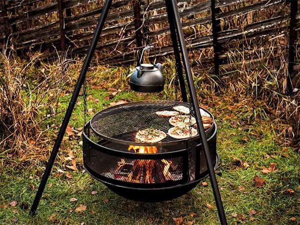 Svart bålgrill på tripod med flammende ild, pizza på rist og hengekjele i høstlig hage.