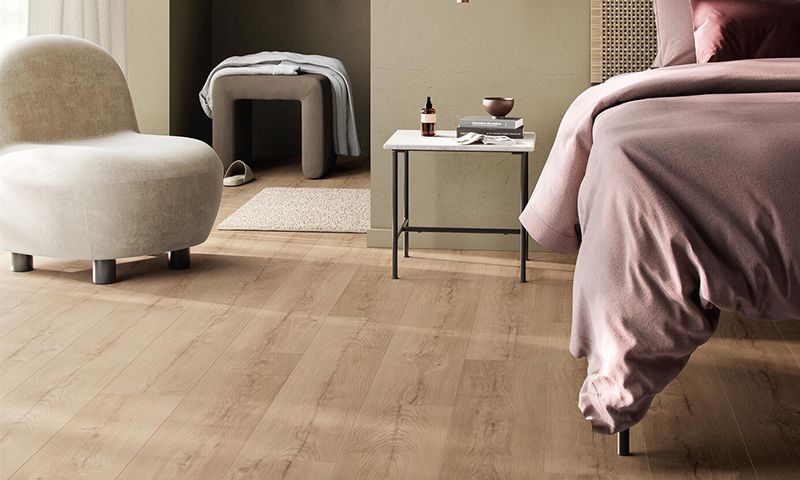 Soverom med dus rosa seng, lyst tregulv, beige lenestol og hvitt nattbord med bok og flaske.