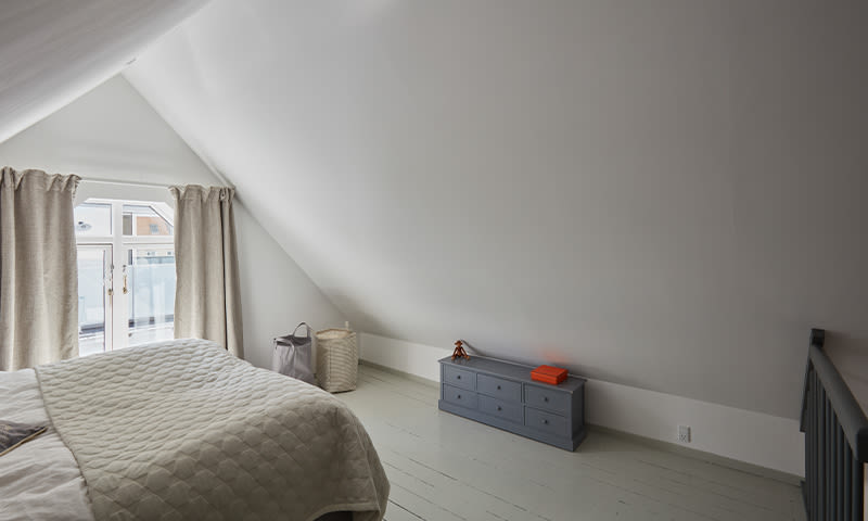 Lyst loftsoverom med skråtak, seng ved vindu med beige gardiner, kurver og blå kommode med rød bok.