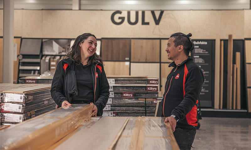 To ansatte i svarte og røde jakker undersøker lyse gulvplanker foran en hylle merket GULV.