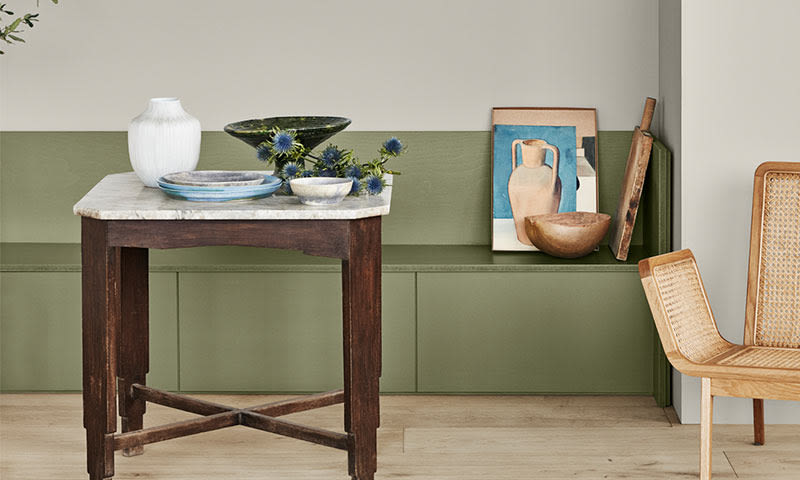 Lite marmorbord med vase, grønne skåler og blå tistler foran olivengrønn skjenk og rottingstol.