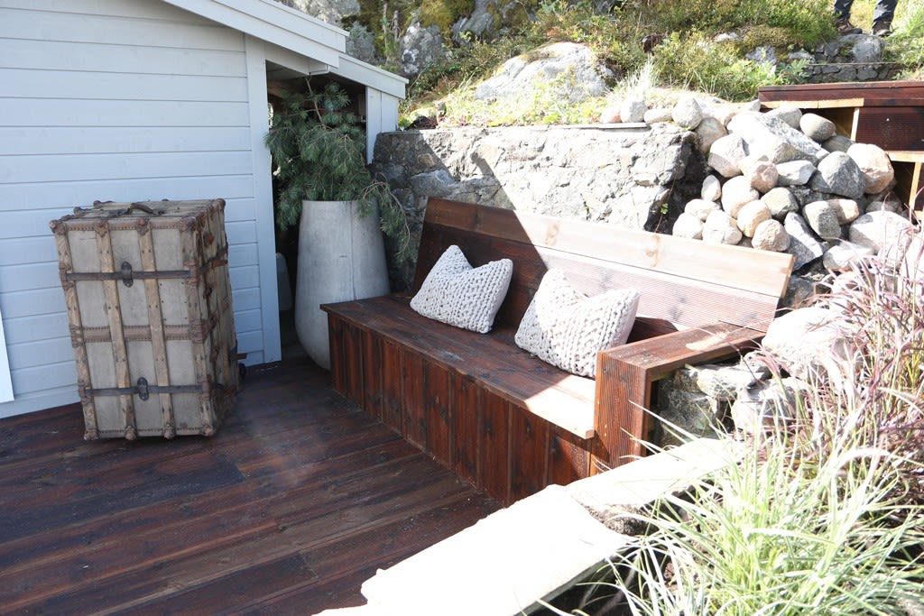 Uteområde med mørkt terrassebord, brun trebenk med hvite strikkputer, vintage koffert, steinmurer.