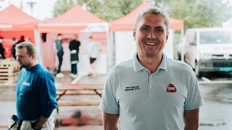 Mann i lys blå BM-polo foran røde messetelt, våte benker og hvit varebil.