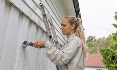 Kvinne maler hvit trepanel på hus fra stige, grønt buskas og rødt tak i bakgrunnen.