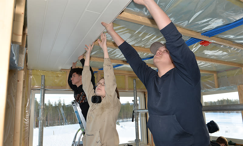 Tre personer løfter hvite takplater mot trebjelker i et uferdig, isolert hjem med snø utenfor.