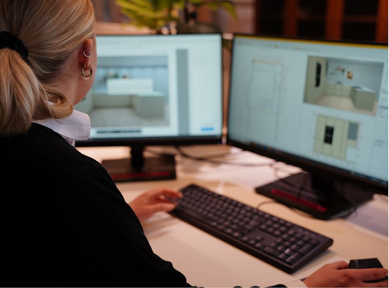 Kvinne ved skrivebord med to skjermer som viser kremhvite kjøkkendesign i programvare.