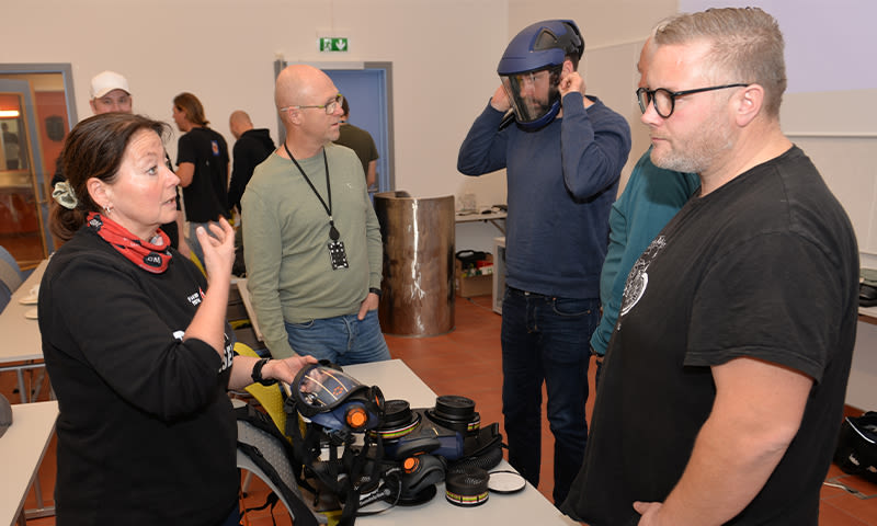 Gruppe ved bord med svarte/gule åndedrettsvernmasker og blå sveisehjelm i lyst rom