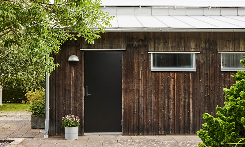 Brun tømmerhytte med svart dør, to smale vinduer og potte med rosa blomster i hage.