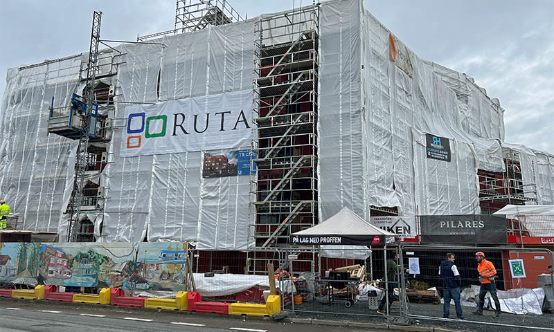 Bygg under hvit byggepresenning med stillas, Ruta-logo, røde/gule sperrebommer og to arbeidere