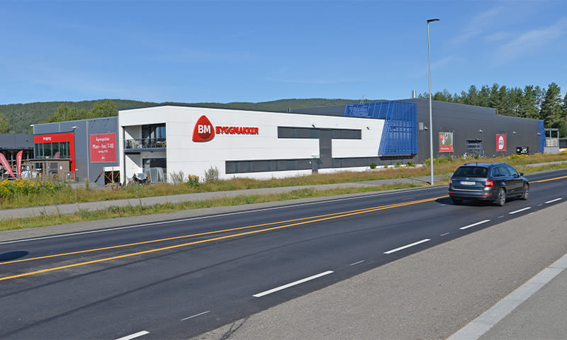 Lang hvitgrå Byggmakker-butikk med rødt logo ved vei, mørk bil under blå himmel