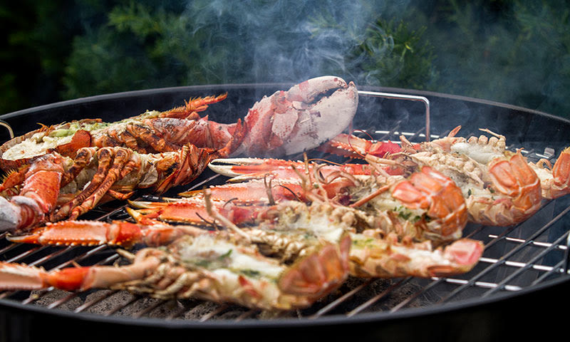 Røde hummerhalvdeler med smør og urter på svart grillrist, grå røyk stiger opp.