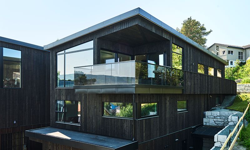 Moderne svart trehus med store vinduer og glassbalkong på grønn skråning