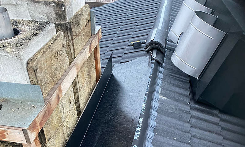 Utsnitt av mørk grå takstein, svart metall-flashing ved åpen skorstein med isolasjon