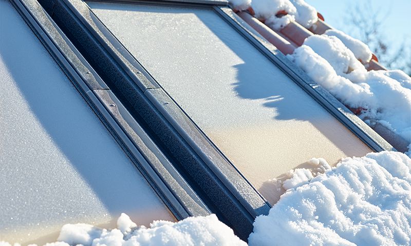 To solcellepaneler på rødt tegltak dekket av snø og rimfrost i solskinn.