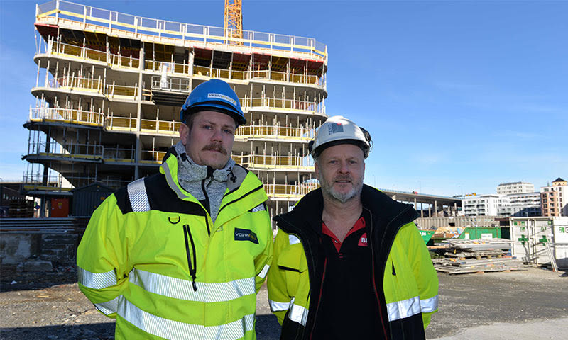 To byggearbeidere i gule vernejakker og hjelmer foran et fleretasje byggeprosjekt og kran.