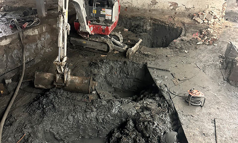 Indre byggeplass med rød-hvitt minigraver, gjørmete gulv og dype firkantede hull.