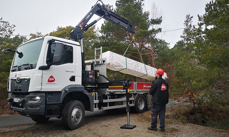 Hvit lastebil med kran løfter hvitpakket bygningslast, arbeider i rød hjelm ved siden av.