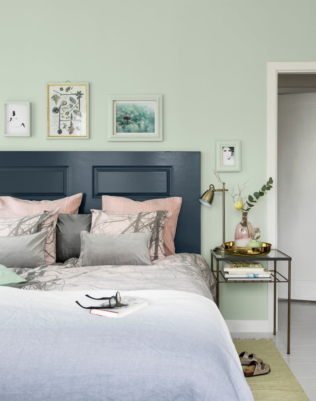 Pastellgrønt soverom med mørkeblå sengegavl, rosa/grå puter, glassbord med messinglampe.