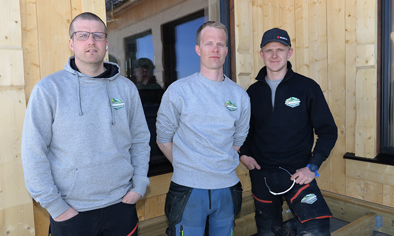 Tre håndverkere i grå og svart arbeidstøy foran trekledning og vinduer på byggeplass.