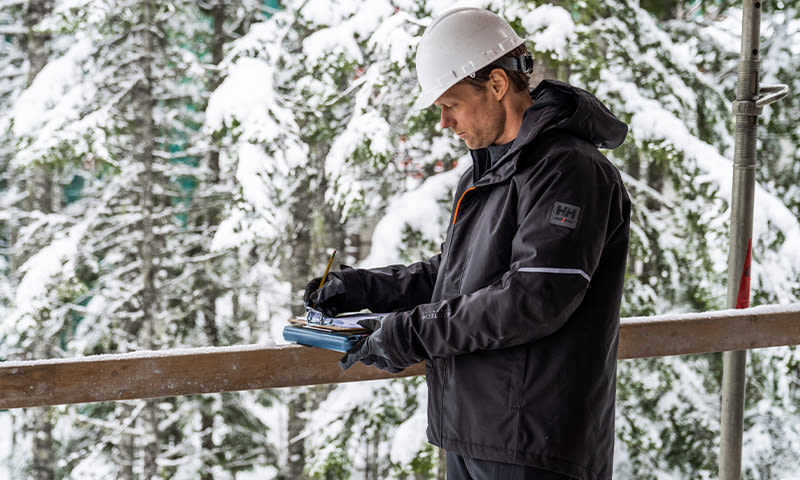 Byggearbeider i svart jakke og hvit hjelm noterer på byggeplass med snødekte trær.