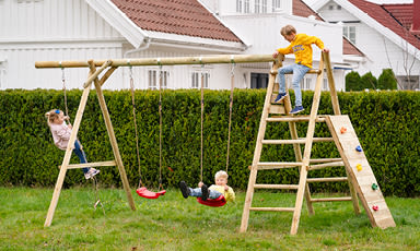 Tre barn leker på lyst treklatrestativ på grønn plen med hvite hus og rødt tak i bakgrunnen