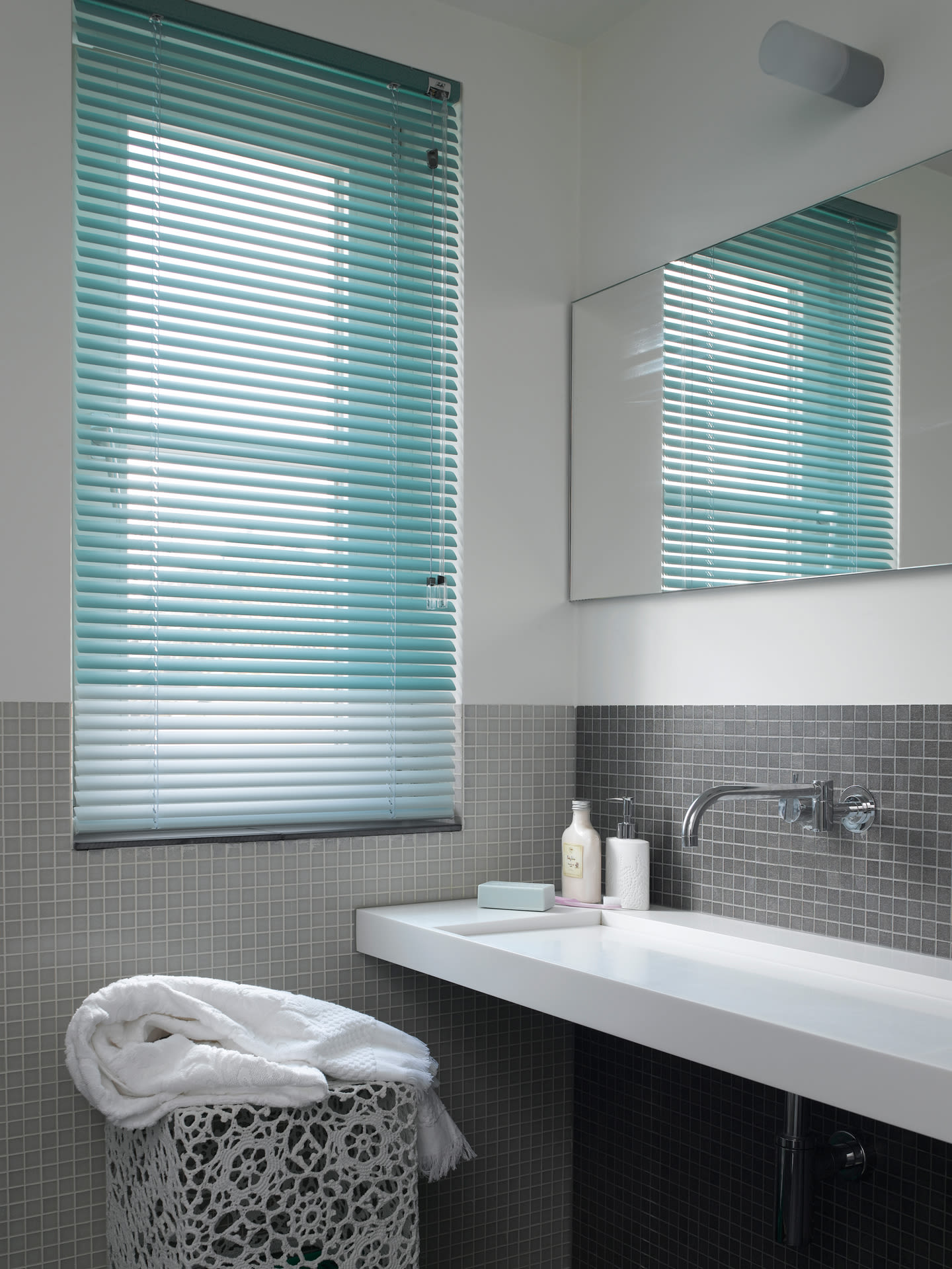 Hvit vask, grå mosaikkfliser, lyseblå persienner, speil og kurv med hvite håndklær.