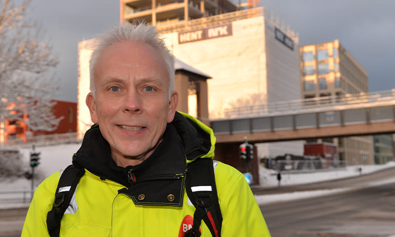 Mann i gul arbeidsjakke med ryggsekk står i snødekt bygate foran kontorbygg og gangbro.