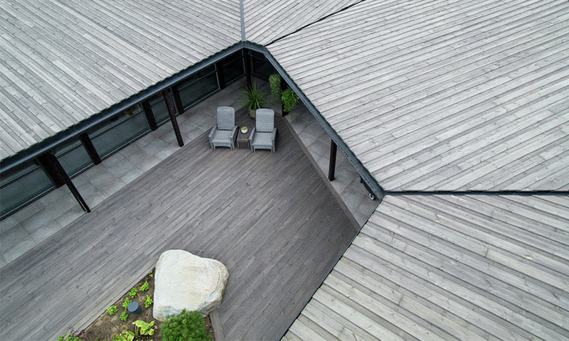 Luftfoto av grå treterrasse omgitt av moderne bygg, med to grå stoler, bord, planter og stor stein