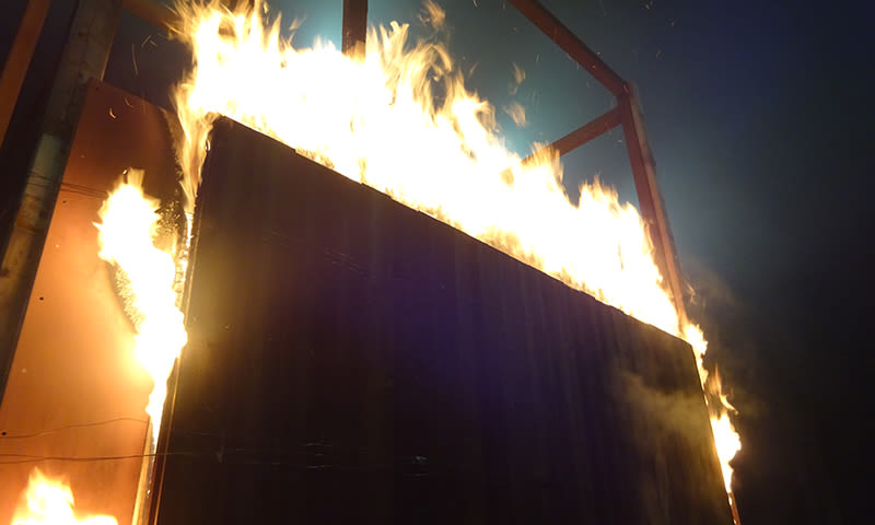 Lysende gule og oransje flammer sluker en mørk fasade mot mørk bakgrunn.