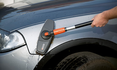 Hånd vasker sølvgrå bil ved hjulbuen med svart-oransje børste og vannperler.