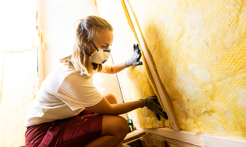 Person i hvit t-skjorte og røde shorts med hansker monterer gul isolasjon i trevegg.