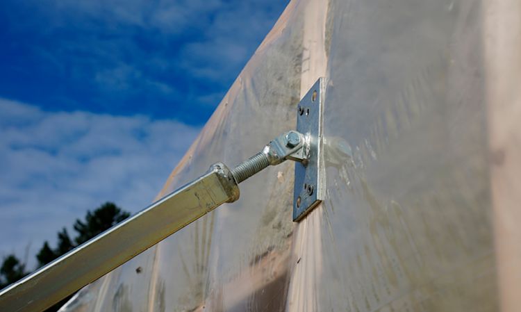 Nærbilde av metallstag med beslag som strammer drivhusplast, blå himmel bak.
