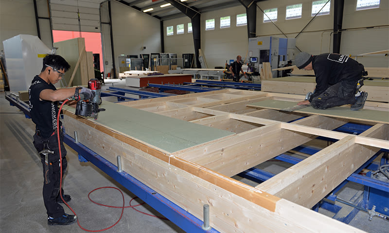 To snekkere bygger store lyse trekonstruksjoner på blå monteringsramme inne i verksted