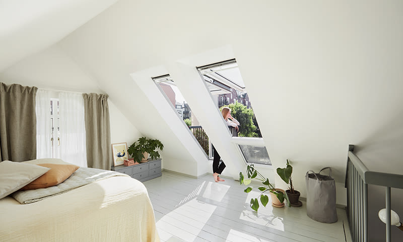 Lyst loftsoverom med skråtak, store skråvinduer, kvinne med stråhatt, beige seng og planter