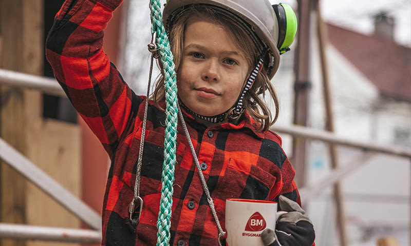 Person i rød-svart rutet skjorte og hvit hjelm med grønne hørselsvern, holder grønt tau og BM-kopp.