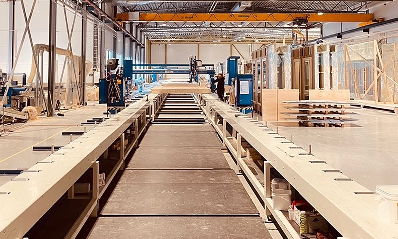 Lys produksjonshall med lange trebånd, blå maskiner, gult traverskran og ansatt.